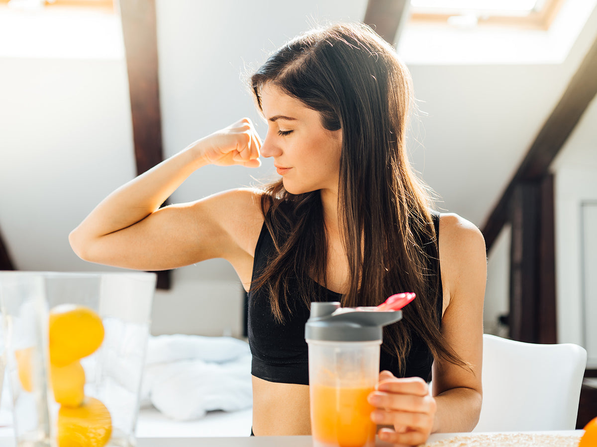 A young woman in workout attire flexes her arm while holding a smoothie, highlighting strength and nourishment for muscles and joints.