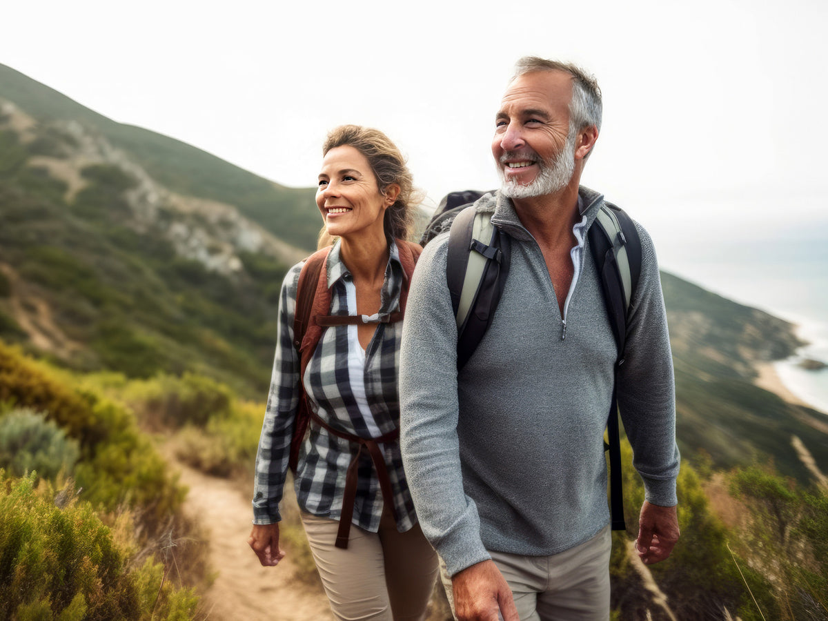 A smiling middle-aged couple hiking through a scenic mountain trail with backpacks, enjoying the outdoors on a bright, clear day.