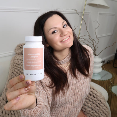 Woman sitting indoors on a beige blanket, smiling and holding a bottle of Hey Nutrition Ashwagandha Complex toward the camera.